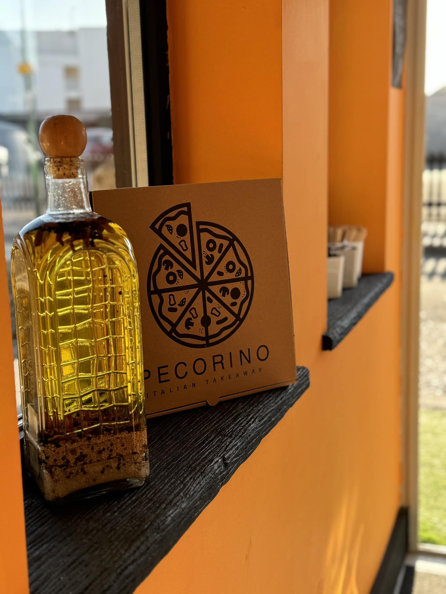 A bottle of olive oil and a pizza box from Pecorino are placed on a shelf against an orange wall